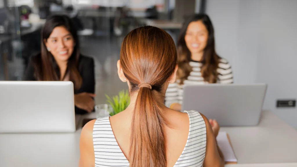 Mujer de espaldas frente a dos entrevistadoras sonrientes con laptops