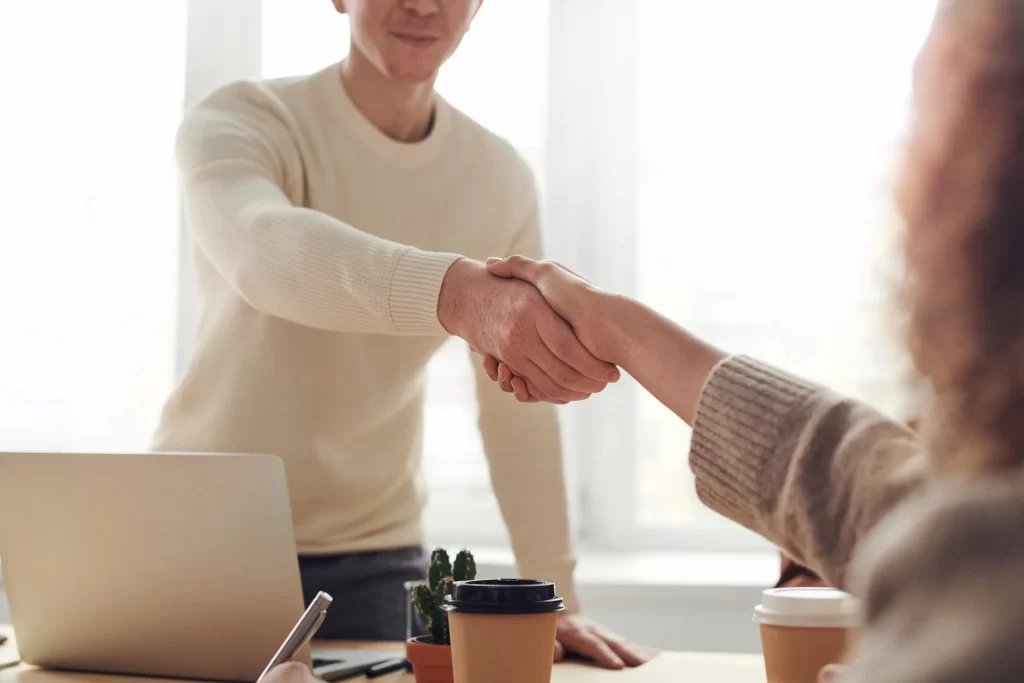 Two people shaking hands over a table with a laptop and coffees